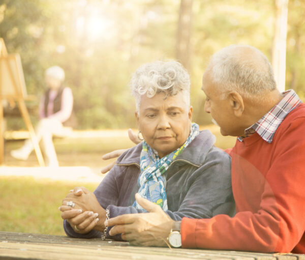 Mixed race senior adult couple talk at outdoor park in spring or autumn season. They sit on a park bench and discuss their relationship difficulties. African and middle eastern descent couple. Woman painting using easel in background.